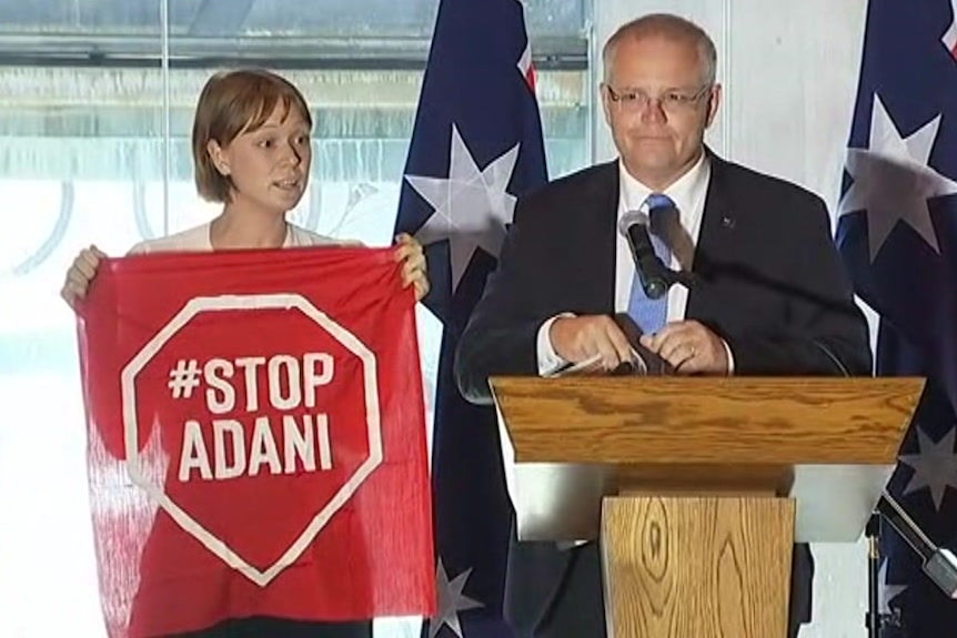 A Stop Adani protester standing next to Scott Morrison on the stage holding a Stop Adani sign.