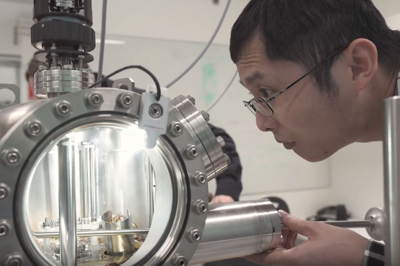 A man peers intently into a metal-and-glass chamber with shiny apparatus inside.