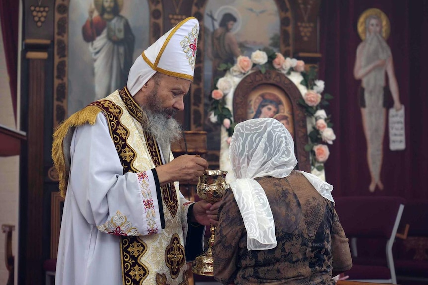 A Coptic church priest delivers a sermon in a church, wearing white robes,with ornate wooden carvings and paintings behind him.