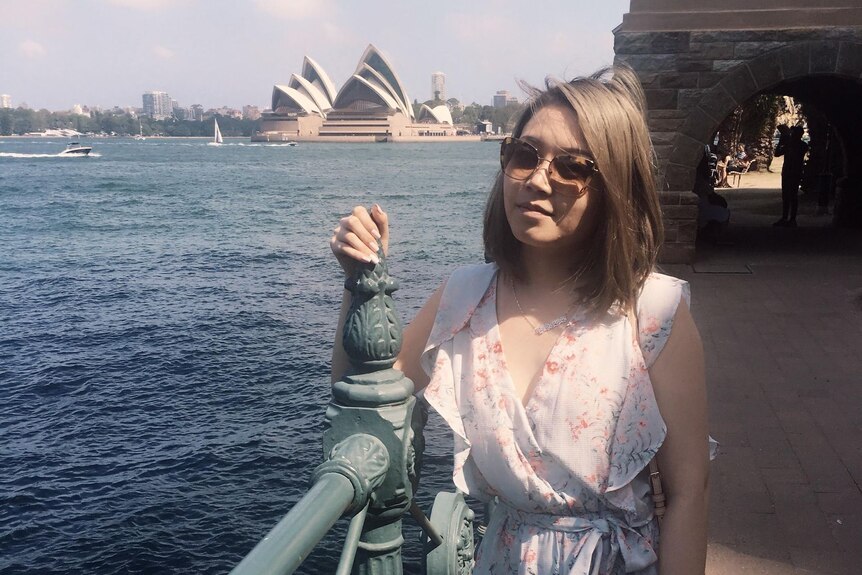 A young Asian woman stands by a railing with the sydney opera house in the background.