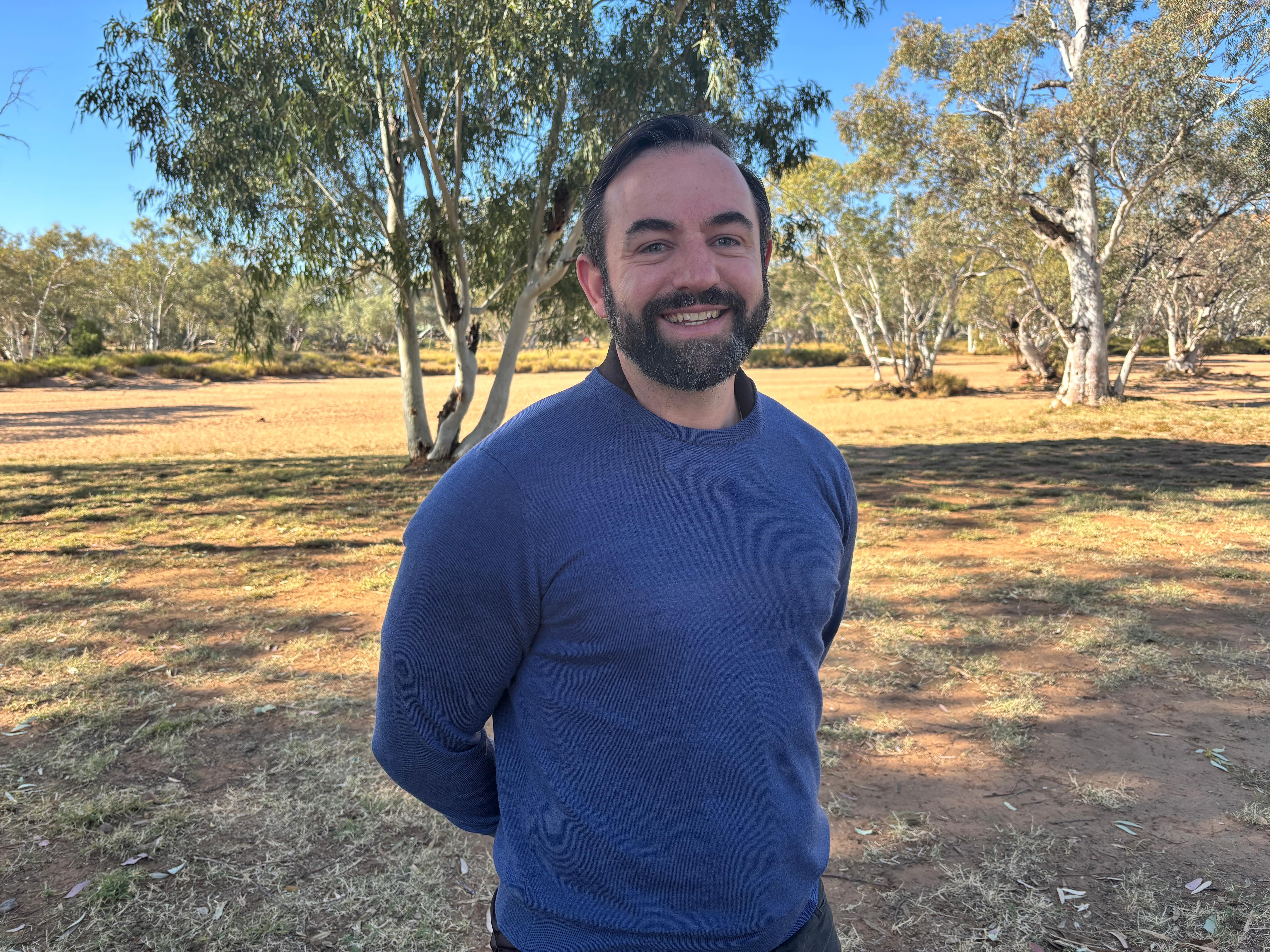 A man in a navy blue jumper stands in front of the Todd River with his hands behind his back and smiles at the camera.