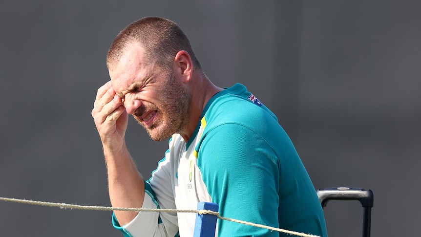 A cricketer in batting pads sits down and squints with his hand on his forehead