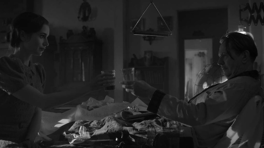 Black and white still with Lily Collins sitting on end of bed and Gary Oldman lying in it, and they're toasting.