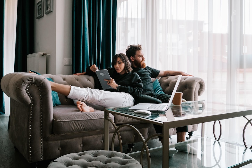 In a light-filled living room a couple sits on a couch. A woman has legs out and leans, reading, on a man, who gazes out window.