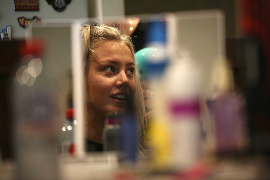 A woman's face can be seen in the reflection of a mirror on a busy table. She is smiling.