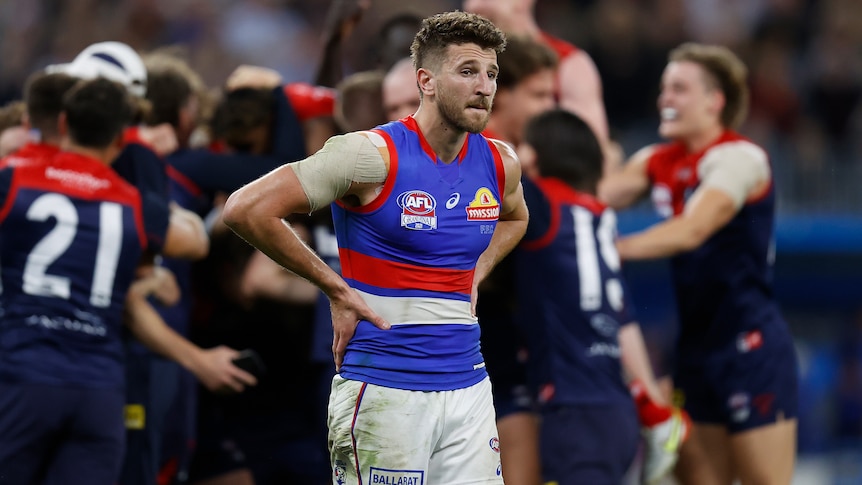 A dejected AFL star stands with hands on hips as opposition players celebrate a grand final win in the background.