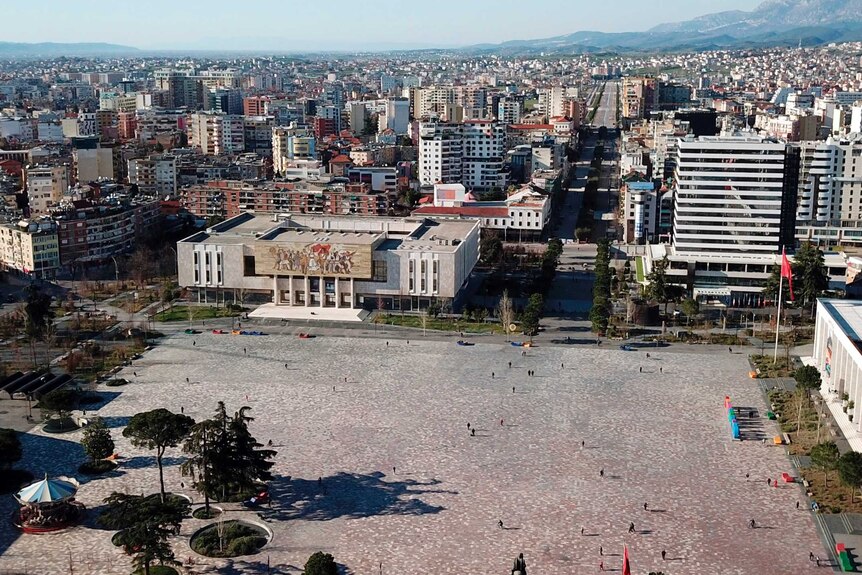 A few people stand in a square, in the Albanian capital of Tirane, with mountains rising in the background.