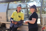 A young man in a high-vis shirt and a woman in a black shirt leaning against a ute tray, talking.