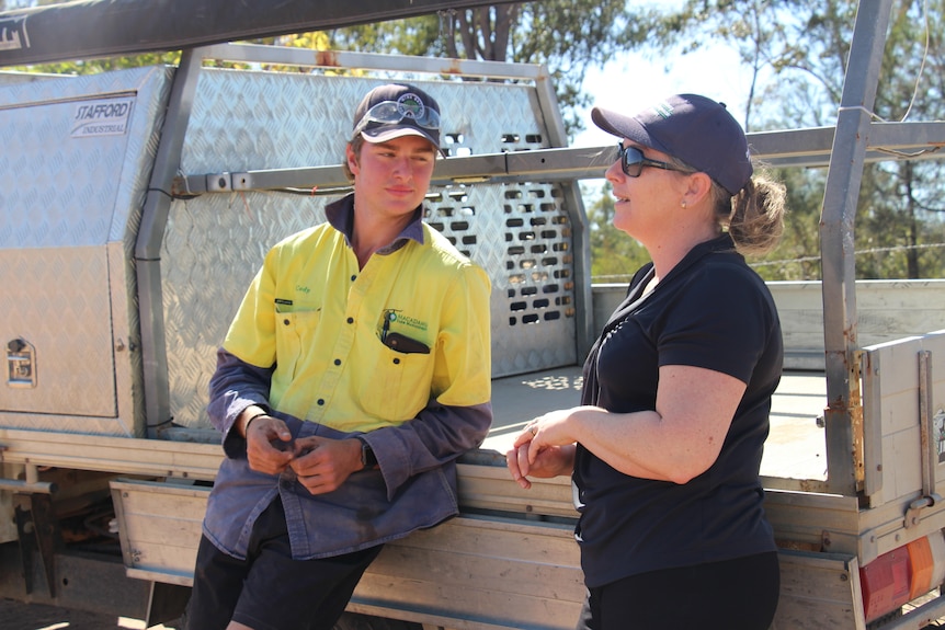 A young man in a high-vis shirt and a woman in a black shirt leaning against a ute tray, talking