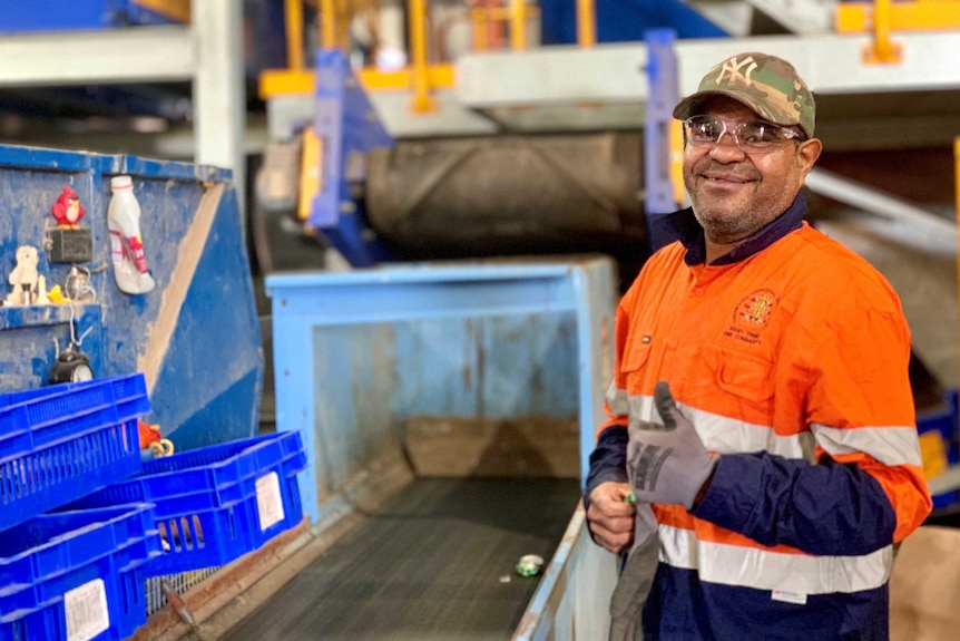 A man smiles at the camera while sorting through recycling materials on a conveyer belt