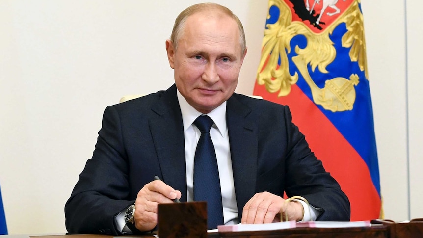 An elderly man with a slight smile sits at a desk in a dark suit with a colourful flag behind him.