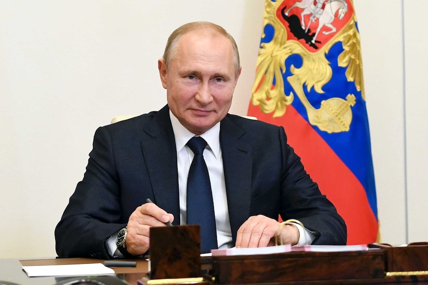 An elderly man with a slight smile sits at a desk in a dark suit with a colourful flag behind him.