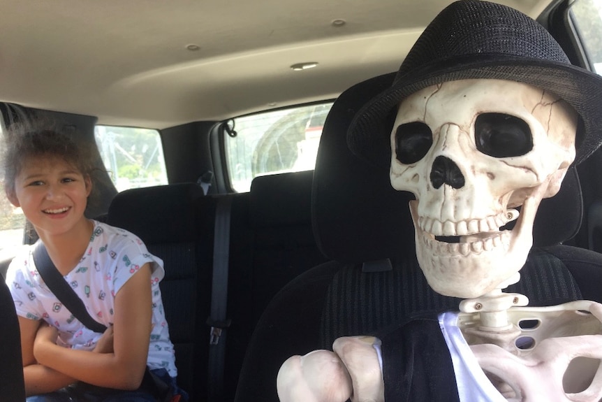 A skeleton wearing a black fedora sits in a car seat with a girl in the back.