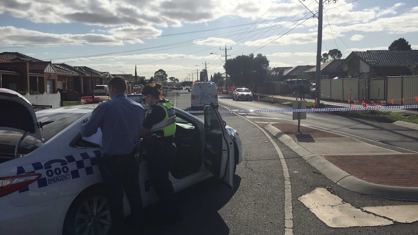 A man in a collared shirt and a policewoman in a high-vis vest stand near a police car and police tape.