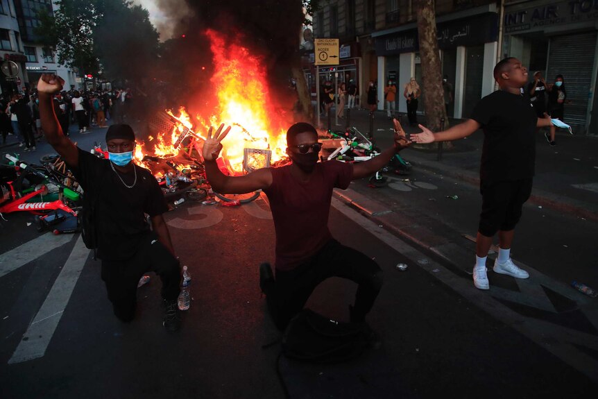 Three protesters kneel in front of a burning barricade with their hands in the air.