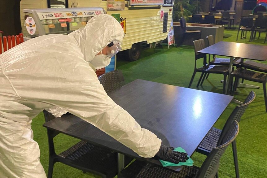 A person dressed in white personal protective gear wipes down a table in front of a caravan.