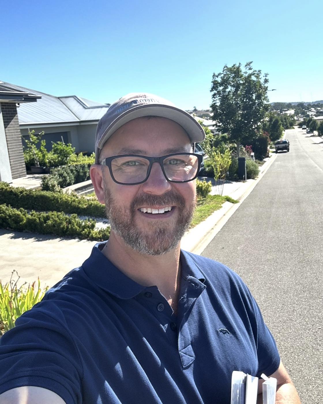 Un hombre con gafas y sombrero se toma un selfie en una calle suburbana en un día soleado