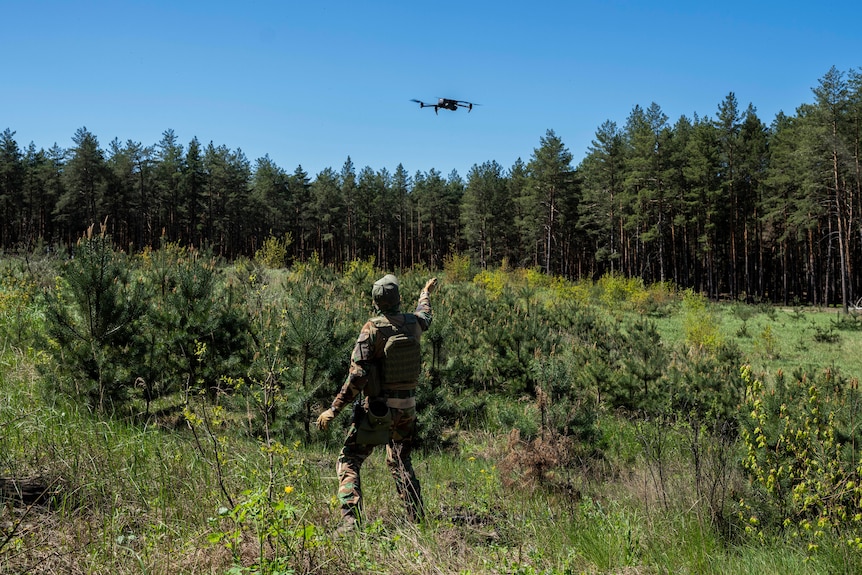 A Ukrainian Border Guard reaches out their hand as a drone hovers above bushland in Ukraine