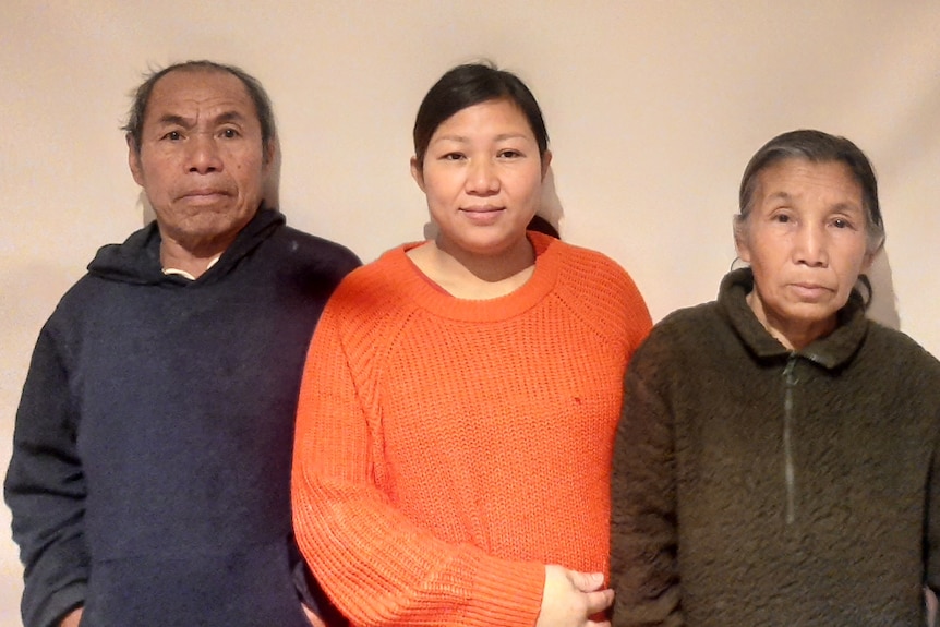 A Karenni woman in orange jumper with her parents standing either side. 
