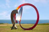 Sculpture made from silver and red steel circular elements, against blue sky and ocean backdrop.