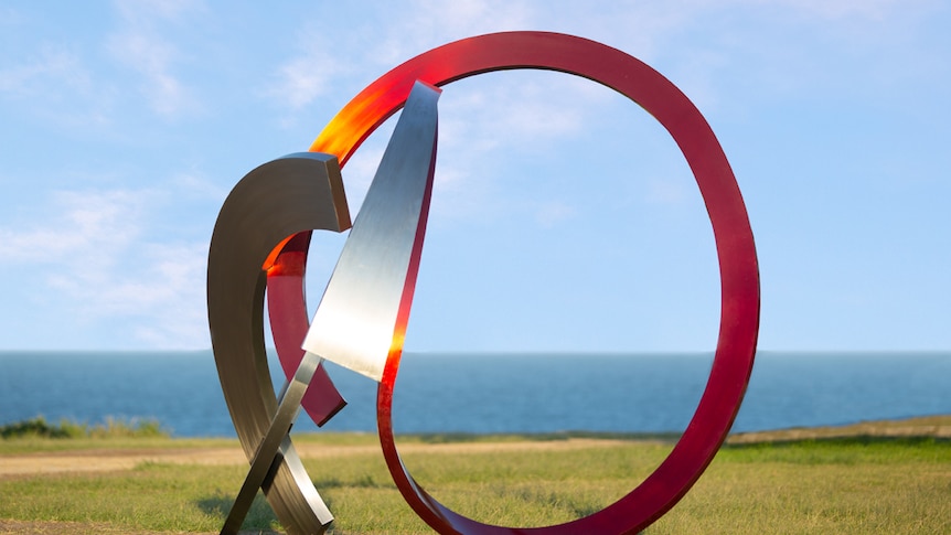 Sculpture made from silver and red steel circular elements, against blue sky and ocean backdrop.