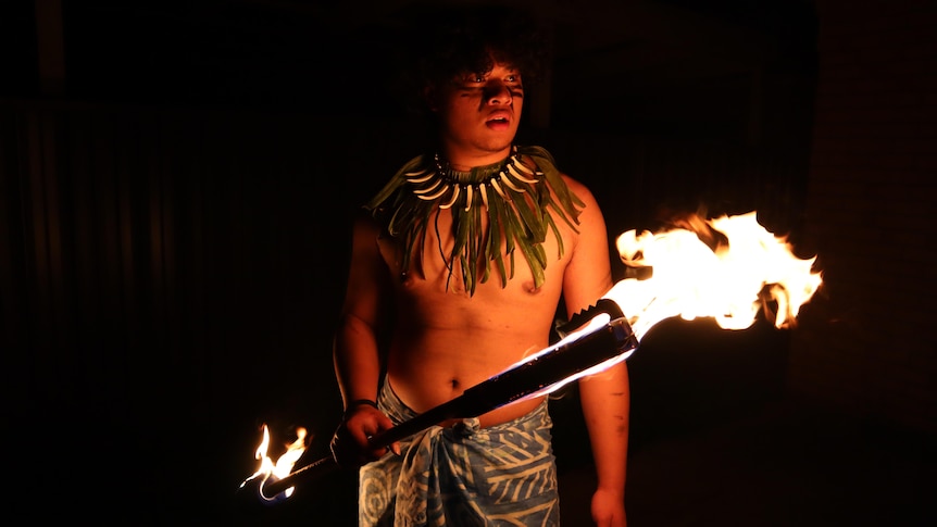 A man in traditional dress stands in darkness illuminated by large flames on either end of a long club.