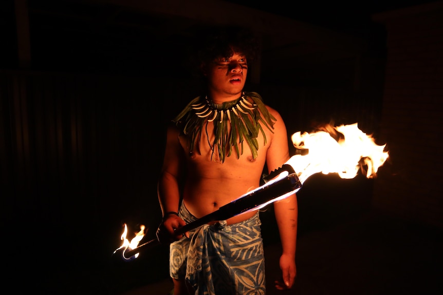 A man in traditional dress stands in darkness illuminated by large flames on either end of a long club.