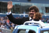 A bearded former AFL player holds a little child and waves to the crowd as he rides in a car around a stadium.