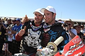 Two men wearing black motocross shirts and white baseball caps stand on a podium