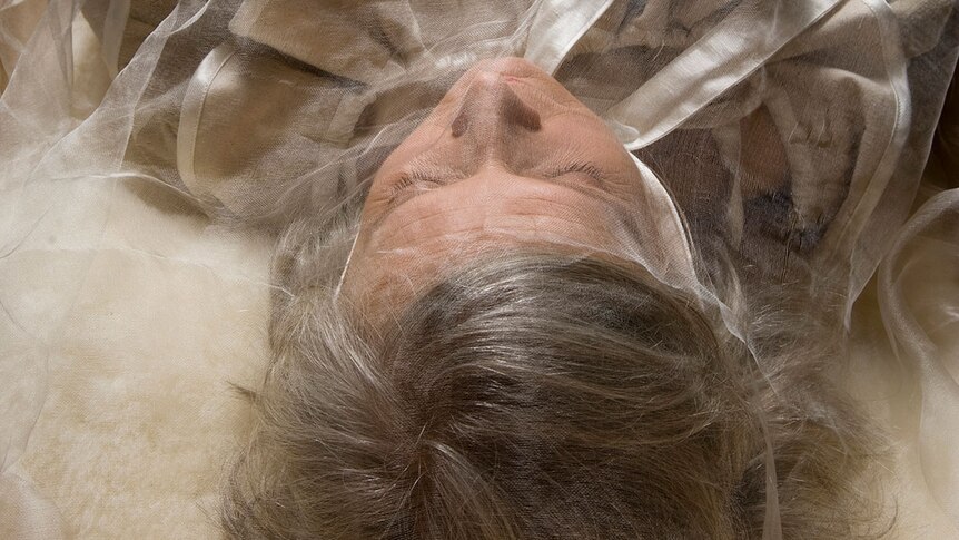 Close up of an older woman's head, covered in a silk shroud, as she lays down.