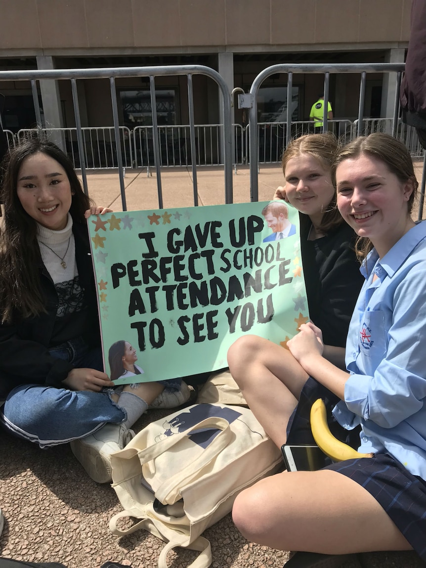 Three female school students sit on the ground with a sign reading "I gave up perfect school attendance to see you"