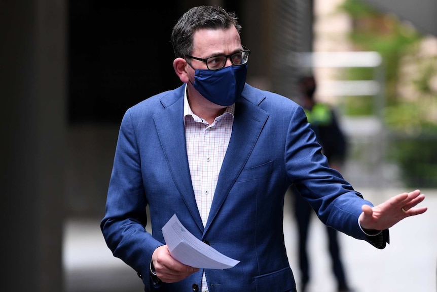 Daniel Andrews, wearing a blue face mask, carries papers in one hand and waves with the other.