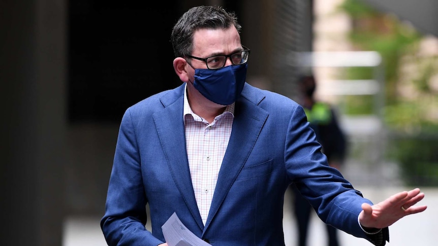 Daniel Andrews, wearing a blue face mask, carries papers in one hand and waves with the other.