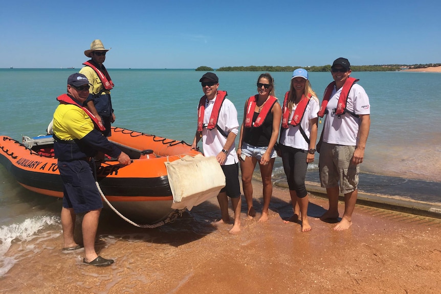 A group of scientists stands next to a boat on the shore as they prepare for a research voyage.