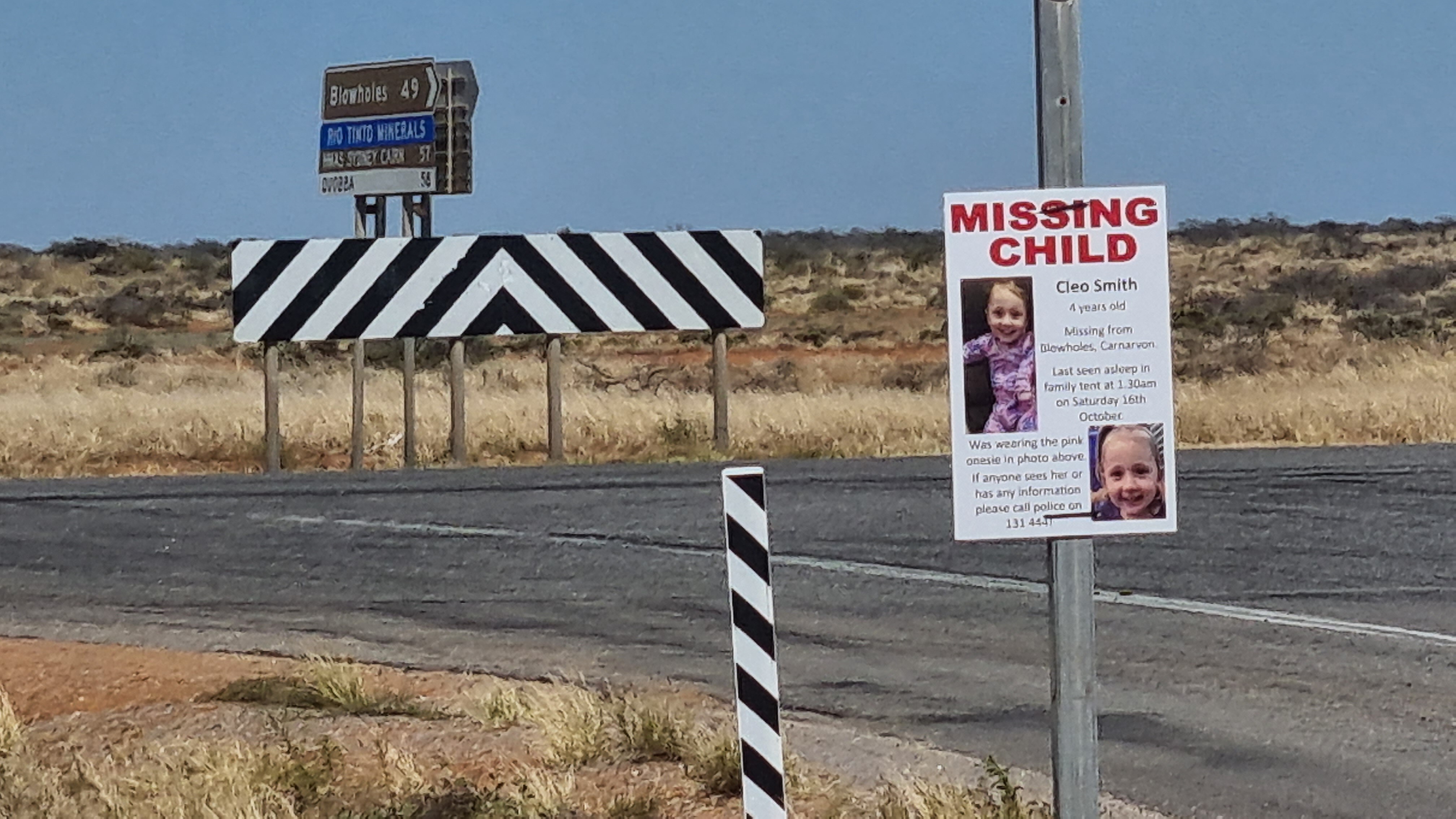 A remote intersection with road signs in the background and a Cleo Smith missing child poster on a pole in the foreground.