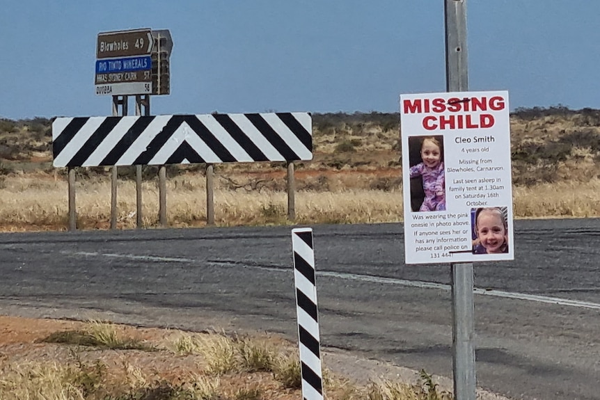 A remote intersection with road signs in the background and a Cleo Smith missing child poster on a pole in the foreground.