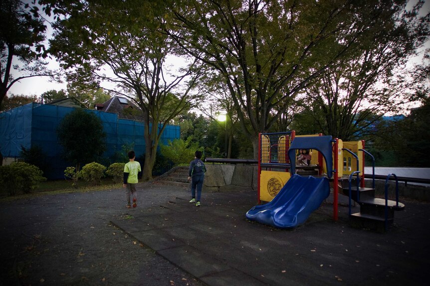Children play on a playground with a house shrouded in blue tarps behind it