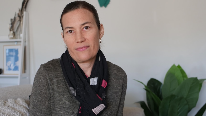 A woman with dark hair wearing a grey top and multi-coloured scarf sits in a loungeroom