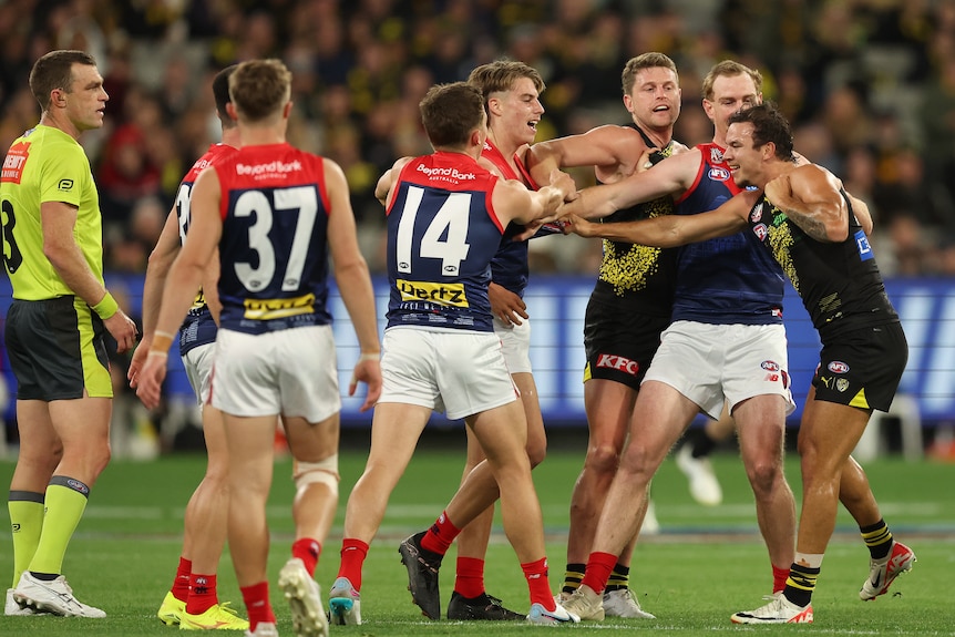 AFL players from Richmond and Melbourne grab each others' guernseys and grapple on the ground as an umpire looks on. 