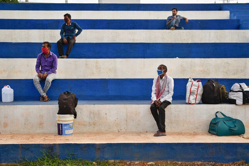 Men sit socially distanced with masks and luggage, on blue and white painted steps.