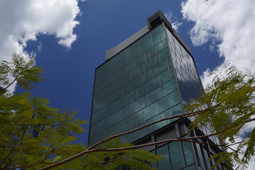 Mirando hacia un edificio alto de la ciudad con ventanas reflectantes azules, con árboles y nubes.