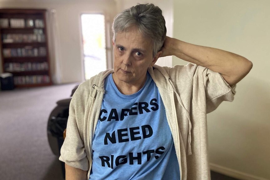 Julie Couzens, wearing a blue t-shirt with 'Carers need rights' on it, stands in a house with her hand behind her head.