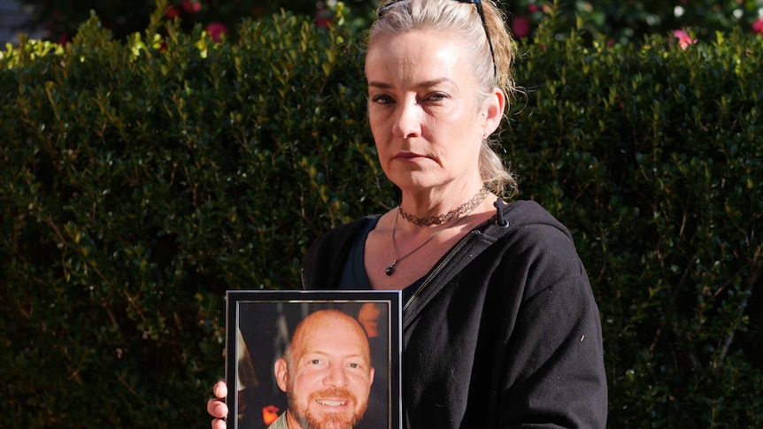 A woman with greying, ash blonde hair holds up a photo of a smiling, bald, bearded man.
