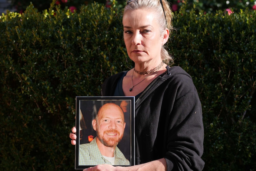 A woman with greying, ash blonde hair holds up a photo of a smiling, bald, bearded man.