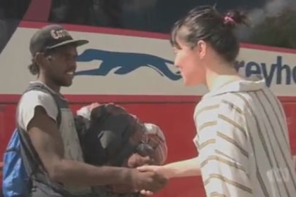 Female journalist shakes hands with an aboriginal youth in front of a bus.