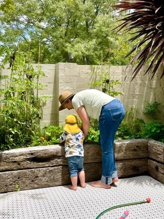 A woman and a child stand with their backs to the camera, looking at a vegetable garden.