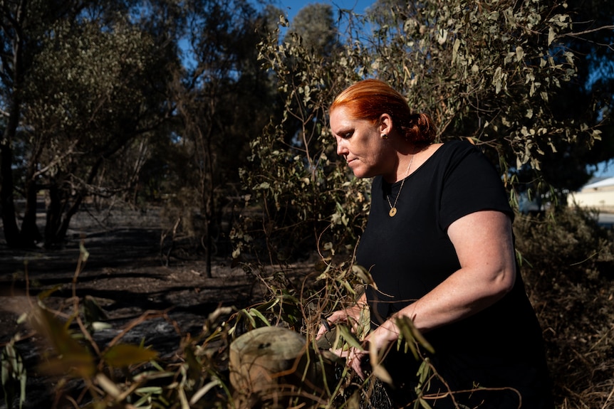 A side-on mid-shot of a middle aged woman wih red hair in the yard of a semi-rural home.