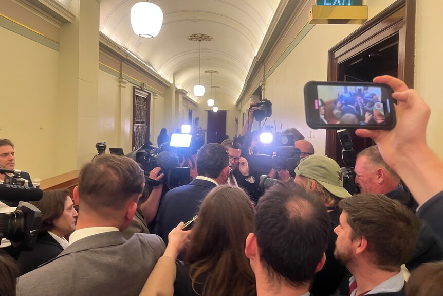The back of Daniel Andrews entering a room surrounded by media with camera's, lights and phones.