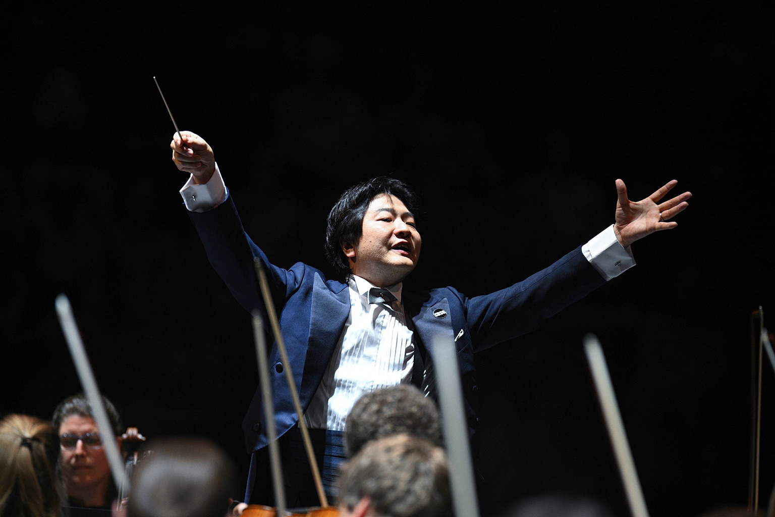 Kazuki Yamada conducts in a blue suit raises baton and arms above an orchestra during a performance on a darkened stage.