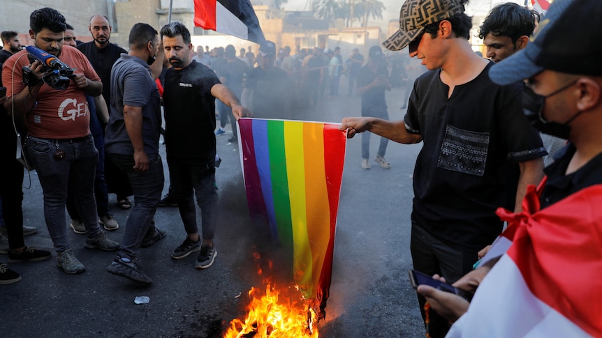 a rainbow flag being lowered into a fire on the floor by two people among a crowd